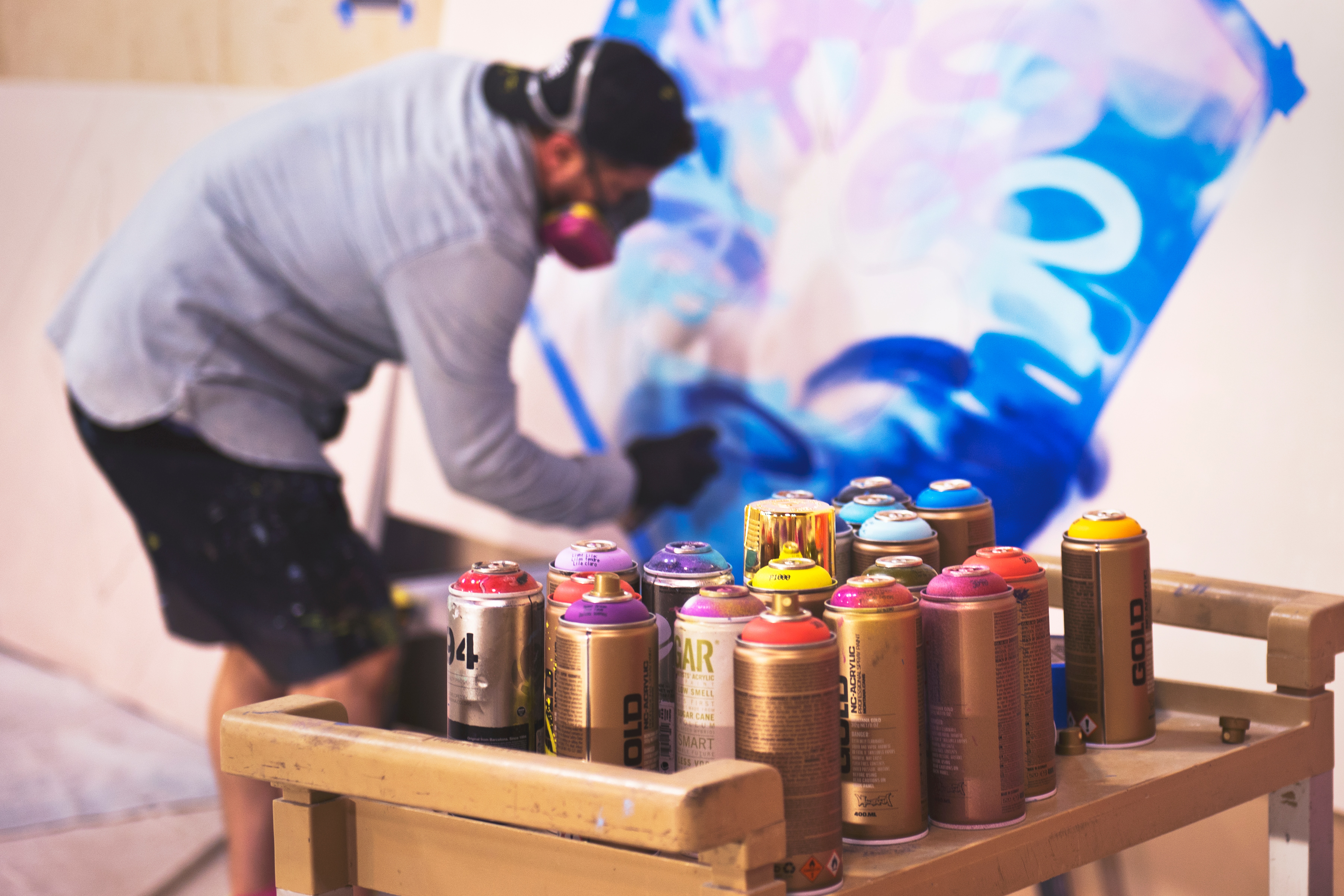Graffiti.jpg Graffiti Spraydosen im Vordergrund. Im Hintergrund eine Person, die ein Graffiti auf eine Leinwand sprüht.